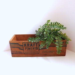 May include: A vintage wooden box with the text "KRAFT PIMENTO" in black lettering. The box is filled with green leafy plants. The box is rectangular and has a rustic, aged appearance, suggesting it is an antique or a repurposed item.