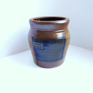 May include: A brown ceramic crock with a wide, rounded rim and a smooth, glossy finish. The front of the vessel features a debossed, dark blue rectangular design depicting a stylized flag with a grid of dots in the upper left corner.