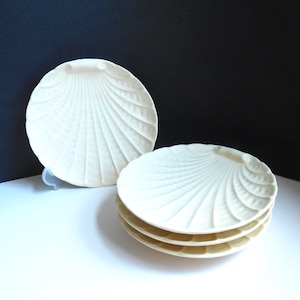 May include: A set of off-white, shell-shaped plates. The plates have a textured, fan-like design radiating from the center. One plate is propped up, while the others are stacked. The background is a gradient of black and white.