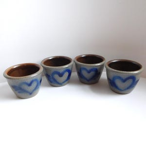 May include: Four small, handmade ceramic bowls. Each bowl is gray with a blue heart design and a brown interior. The bowls are arranged on a white surface, showcasing their rustic charm and unique craftsmanship.