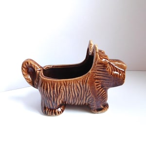May include: A brown ceramic planter shaped like a Scottish Terrier dog. The piece features a glossy finish with molded fur texture details, a curled tail, and an open top cavity for holding small items or plants, standing on four short legs.