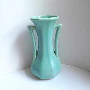 May include: A light turquoise ceramic vase with a smooth, matte finish. The vessel features a hexagonal body, a flared rim, and two symmetrical, curved handles extending from the neck to the shoulder, creating a balanced and structured silhouette.