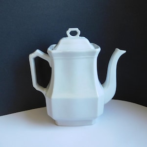 May include: A white porcelain teapot with a lid, handle, and spout. The teapot has a geometric design with a square base and a rectangular body. The handle is a square shape, and the spout is curved. The lid has a small handle.