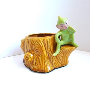 May include: A ceramic planter shaped like a brown tree stump with a textured bark finish. A small, light green elf figure with a pointed hat and painted facial features is perched on the edge of the stump, resting one arm on its knee.