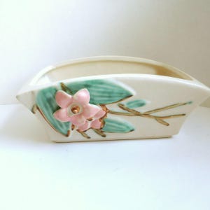 May include: Cream-colored ceramic planter with a curved handle. The planter is decorated with a pink flower and teal leaves, with gold accents on the flower's center and the branches. The planter has a rectangular shape.