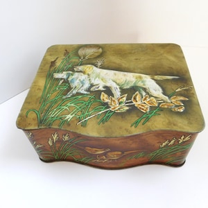 May include: A vintage, square-shaped metal box with a hinged lid. The lid features a painted scene of two dogs in a field with tall grass and foliage. The box has a brown base with painted details of plants and birds.