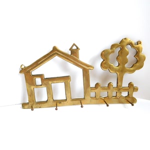 May include: A brass key holder shaped like a house, tree, and fence. The key holder has a chimney, windows, and a tree with a rounded top. It features five hooks for keys and is made of gold-colored metal.
