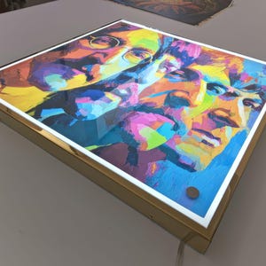 Peut inclure: Impression d'art carrée avec un portrait abstrait coloré. L'œuvre d'art a un cadre doré avec une bordure blanche. Le portrait est composé de bleus, de jaunes, de roses et d'oranges vifs. Dimensions: environ 30 cm x 30 cm.