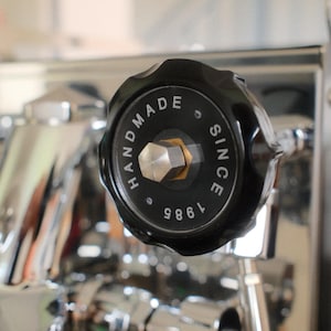 May include: Close-up of a black knob with the text "HANDMADE SINCE 1985" in white lettering. The knob is part of a shiny, chrome-colored espresso machine. The knob has a hexagonal metal piece in the center.