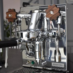 Puede incluir: Una máquina de espresso cromada con pomos de madera y cuerpo de acero inoxidable. La máquina tiene un portafiltro simple y una varilla de vapor. La máquina está etiquetada como "PROFITEc".