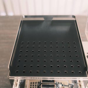 Peut inclure: Gros plan sur une machine à expresso en acier inoxydable avec un bac d'égouttage noir. Le bac comporte une grille de petits trous carrés. La machine a une surface polie et réfléchissante et est posée sur une surface claire.