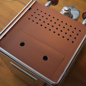 Könnte beinhalten: Nahaufnahme einer Espresso-Maschine aus Edelstahl und Kupfer. Die Oberseite hat eine kupferfarbene Oberfläche mit mehreren kleinen Löchern. Die Maschine hat Knöpfe aus Holz und einen silbernen Griff. Das Design ist modern und industriell.