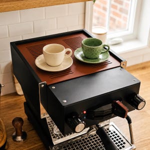 Può includere: Una macchina per caffè espresso nera con un vassoio superiore color ruggine. Due tazze in ceramica con piattini abbinati sono posizionate sopra: una tazza color crema tinta unita e una tazza verde chiaro maculata. La macchina ha manopole nere.