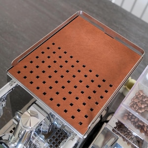 May include: A rectangular espresso machine cup warming tray featuring a smooth, brown, leather-like surface with a grid of square ventilation cutouts on the left side and a solid section on the right, framed by a polished, reflective metal border.