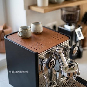 Puede incluir: Una máquina de café espresso negra con un panel superior texturizado de color marrón que presenta dos secciones cuadradas perforadas. Dos tazas pequeñas de cerámica color hueso descansan sobre la superficie. La máquina tiene un frente cromado.