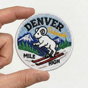 May include: Embroidered patch featuring a white ram skiing down a snowy slope. The patch is round with a light blue background and the words "DENVER Colorado" and "MILE HIGH" in black lettering. Green trees and mountains are also depicted.
