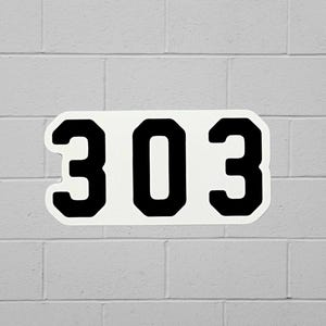 May include: A white rectangular sign with the number "303" in bold black font. The sign has rounded corners and is set against a gray brick wall.