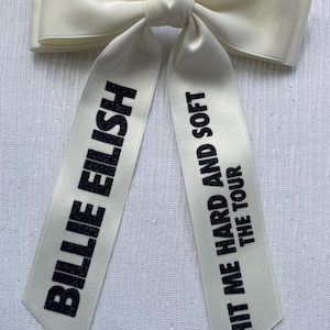 May include: A cream-colored satin hair bow with two long tails. The left tail features the black glitter text BILLIE EILISH, and the right tail features the black glitter text HIT ME HARD AND SOFT THE TOUR, printed in a bold, sans-serif font.