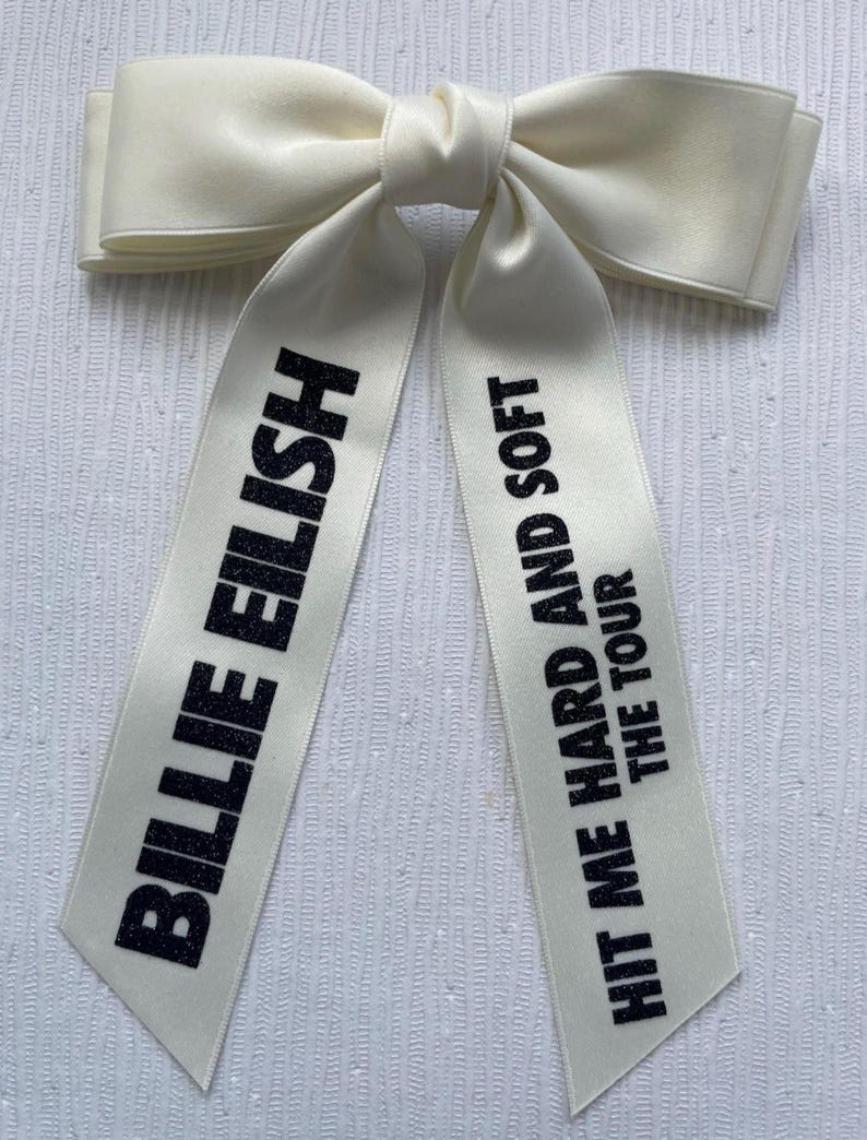 May include: A cream-colored satin hair bow with two long tails. The left tail features the black glitter text BILLIE EILISH, and the right tail features the black glitter text HIT ME HARD AND SOFT THE TOUR, printed in a bold, sans-serif font.