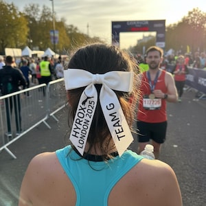 May include: A white hair bow with two long ribbons attached, featuring the black printed text HYROX LONDON 2025 and the Puma logo on one side, and the text TEAM G on the other side, worn in dark hair above a bright turquoise racerback tank top.