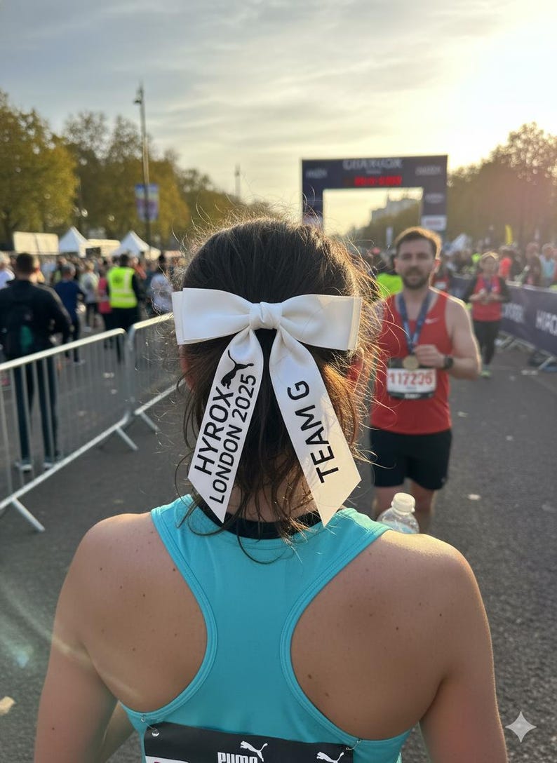 May include: A white hair bow with two long ribbons attached, featuring the black printed text HYROX LONDON 2025 and the Puma logo on one side, and the text TEAM G on the other side, worn in dark hair above a bright turquoise racerback tank top.