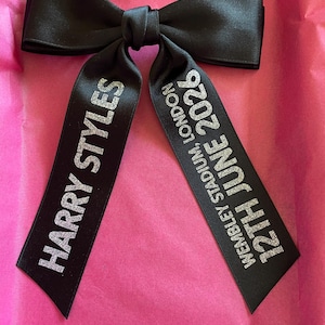 May include: A black satin hair bow featuring silver glitter text. The left tail reads HARRY STYLES, and the right tail reads WEMBLEY STADIUM, LONDON 12TH JUNE 2026. The bow is constructed with a central knot and two long, hanging ribbon tails.