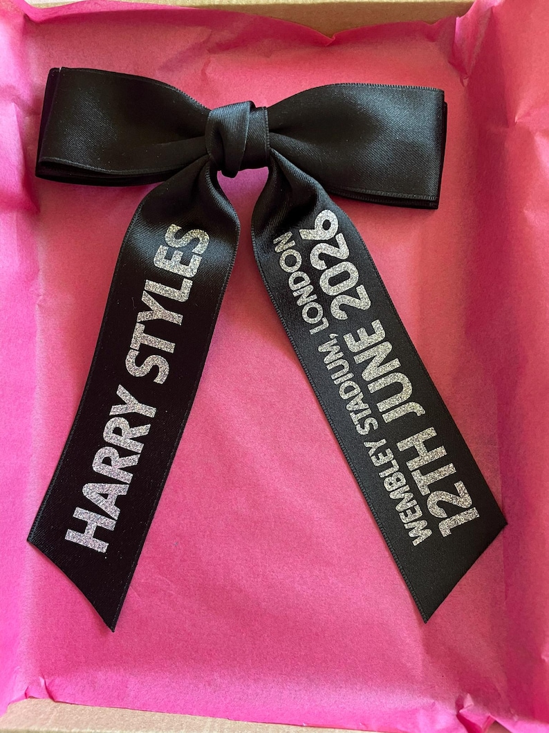 May include: A black satin hair bow featuring silver glitter text. The left tail reads HARRY STYLES, and the right tail reads WEMBLEY STADIUM, LONDON 12TH JUNE 2026. The bow is constructed with a central knot and two long, hanging ribbon tails.