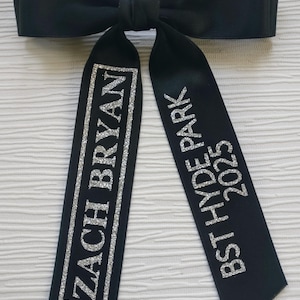 May include: A black satin hair bow with two long tails. The left tail features the silver glitter text ZACH BRYAN enclosed in a rectangular border. The right tail features the silver glitter text BST HYDE PARK 2025 stacked vertically.