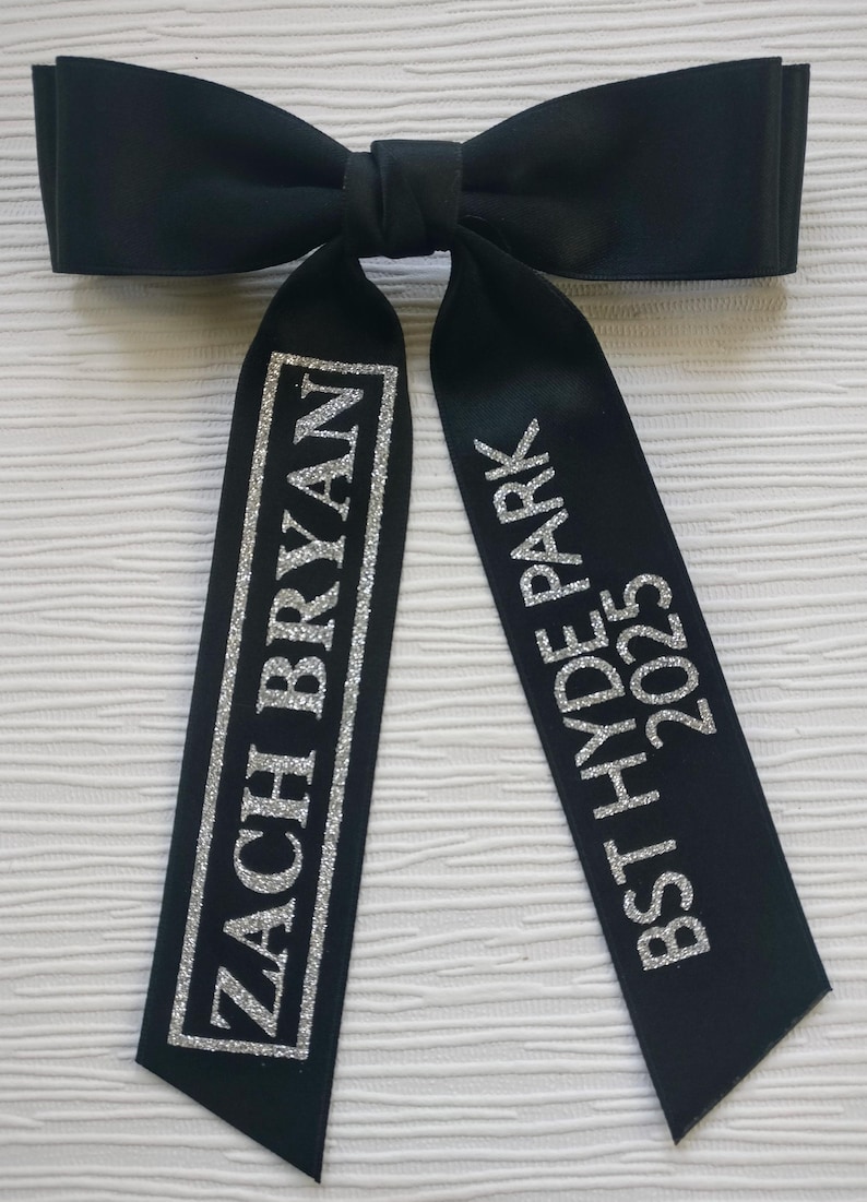 May include: A black satin hair bow with two long tails. The left tail features the silver glitter text ZACH BRYAN enclosed in a rectangular border. The right tail features the silver glitter text BST HYDE PARK 2025 stacked vertically.