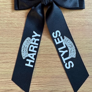 May include: A black satin hair bow with two long, hanging tails. The left tail features the white text HARRY with the word TOGETHER printed above it, and the right tail features the white text STYLES with the word TOGETHER printed above it.