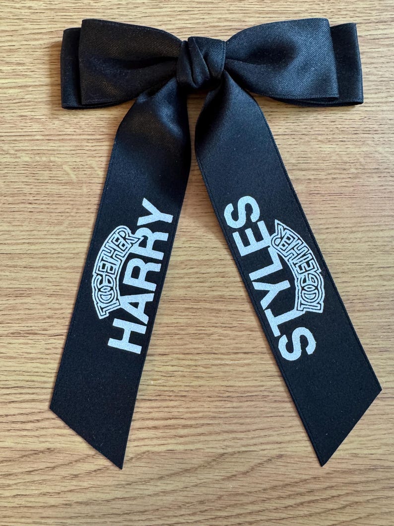 May include: A black satin hair bow with two long, hanging tails. The left tail features the white text HARRY with the word TOGETHER printed above it, and the right tail features the white text STYLES with the word TOGETHER printed above it.