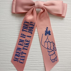 May include: A light pink satin hair bow with two long tails. The left tail features the blue glitter text "SCREW EM IF THEY CAN'T TAKE A JOKE!" and the right tail features a blue glitter graphic of a person wearing a ruffled dress.
