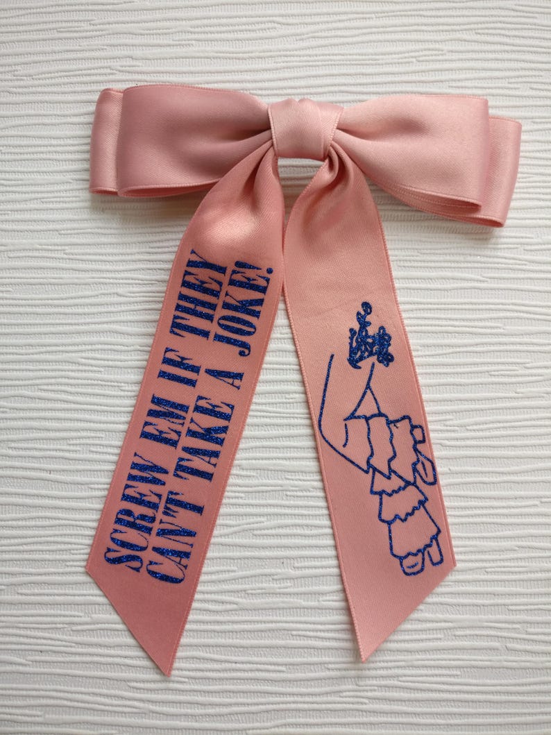 May include: A light pink satin hair bow with two long tails. The left tail features the blue glitter text "SCREW EM IF THEY CAN'T TAKE A JOKE!" and the right tail features a blue glitter graphic of a person wearing a ruffled dress.
