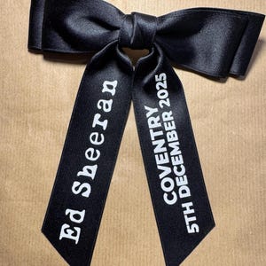 May include: A black satin hair bow with two long tails. The left tail features the white text Ed Sheeran, and the right tail features the white text COVENTRY 5TH DECEMBER 2025. The bow has a smooth, reflective finish and a central knot detail.