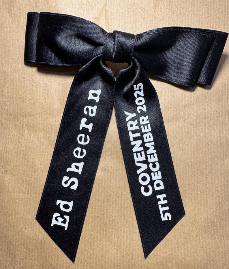 May include: A black satin hair bow with two long tails. The left tail features the white text Ed Sheeran, and the right tail features the white text COVENTRY 5TH DECEMBER 2025. The bow has a smooth, reflective finish and a central knot detail.