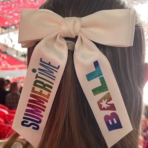 May include: A cream-colored satin hair bow featuring two long, hanging ribbons. The left ribbon displays the word "SUMMERTIME" in a vertical, rainbow-colored gradient, and the right ribbon displays the word "BALL" in the same rainbow gradient.