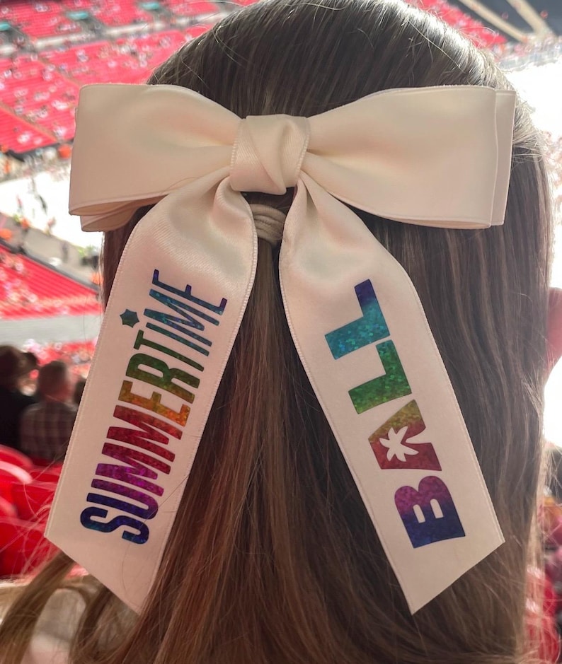 May include: A cream-colored satin hair bow featuring two long, hanging ribbons. The left ribbon displays the word "SUMMERTIME" in a vertical, rainbow-colored gradient, and the right ribbon displays the word "BALL" in the same rainbow gradient.