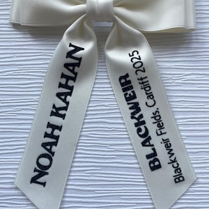 May include: A cream-colored satin hair bow with two long tails. The left tail features the black text NOAH KAHAN, and the right tail features the black text BLACKWEIR Blackweir Fields, Cardiff 2025. The bow has a smooth, shiny finish.