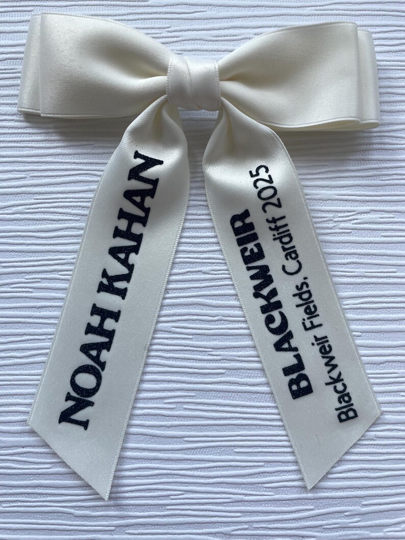 May include: A cream-colored satin hair bow with two long tails. The left tail features the black text NOAH KAHAN, and the right tail features the black text BLACKWEIR Blackweir Fields, Cardiff 2025. The bow has a smooth, shiny finish.
