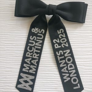 May include: A black satin hair bow with two long tails. The left tail features the silver glitter text MARCUS & MARTINUS with a logo, and the right tail features the silver glitter text WANTS P2 LONDON 2025. The bow has a smooth, matte finish.