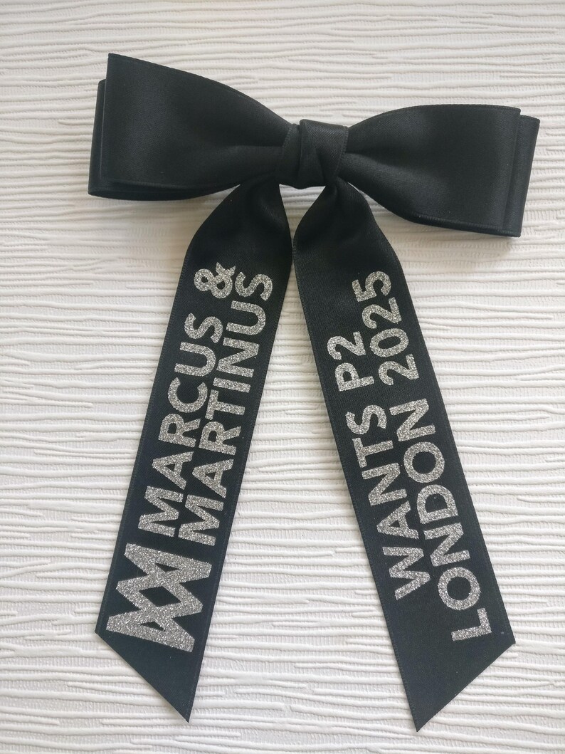 May include: A black satin hair bow with two long tails. The left tail features the silver glitter text MARCUS & MARTINUS with a logo, and the right tail features the silver glitter text WANTS P2 LONDON 2025. The bow has a smooth, matte finish.