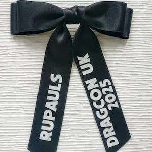 May include: A black satin hair bow featuring two long, hanging ribbons. The left ribbon displays the white text RUPAULS, while the right ribbon features the white text DRAGCON UK 2025. The bow has a smooth, reflective finish and a knotted center.