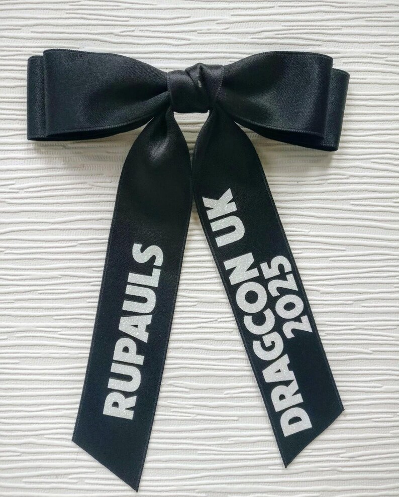 May include: A black satin hair bow featuring two long, hanging ribbons. The left ribbon displays the white text RUPAULS, while the right ribbon features the white text DRAGCON UK 2025. The bow has a smooth, reflective finish and a knotted center.