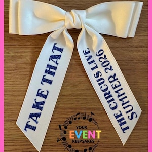 May include: A white satin hair bow with two long tails. The left tail features the blue glitter text "TAKE THAT" and the right tail features the blue glitter text "THE CIRCUS LIVE SUMMER 2026". The bow has a smooth, shiny finish and a knotted center.