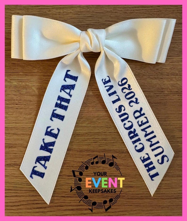 May include: A white satin hair bow with two long tails. The left tail features the blue glitter text "TAKE THAT" and the right tail features the blue glitter text "THE CIRCUS LIVE SUMMER 2026". The bow has a smooth, shiny finish and a knotted center.