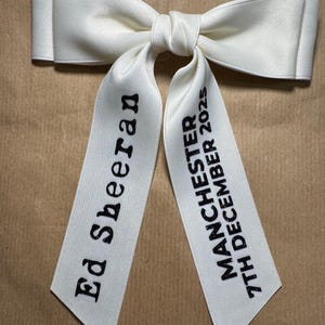 May include: A white satin hair bow with two long tails. The left tail features the black text Ed Sheeran, and the right tail features the black text MANCHESTER 7TH DECEMBER 2025. The bow has a smooth, slightly reflective finish and a central knot.