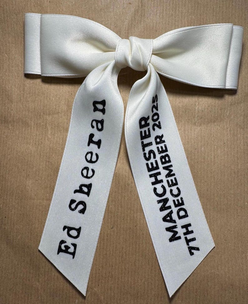 May include: A white satin hair bow with two long tails. The left tail features the black text Ed Sheeran, and the right tail features the black text MANCHESTER 7TH DECEMBER 2025. The bow has a smooth, slightly reflective finish and a central knot.
