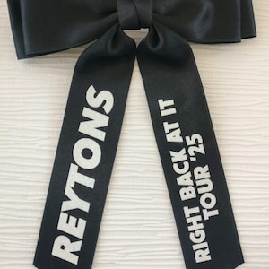 May include: A black satin hair bow featuring two long tails. The left tail displays the white text REYTONS, while the right tail features the white text RIGHT BACK AT IT TOUR '25. The bow has a smooth, slightly reflective finish and a knotted center.