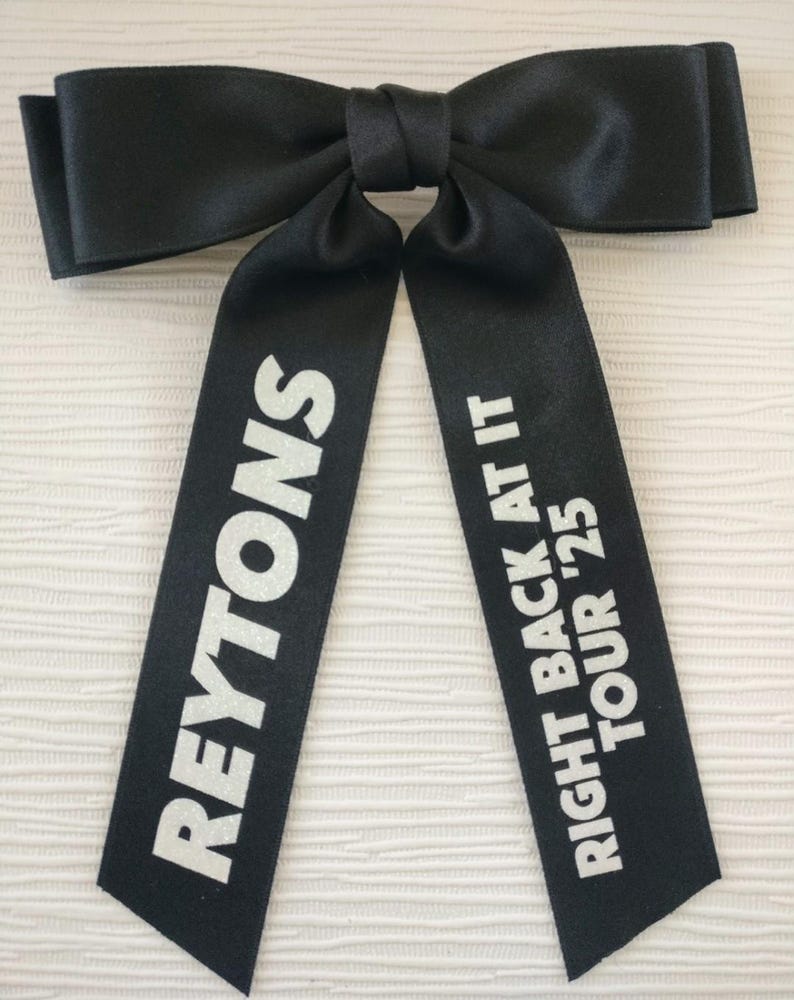 May include: A black satin hair bow featuring two long tails. The left tail displays the white text REYTONS, while the right tail features the white text RIGHT BACK AT IT TOUR '25. The bow has a smooth, slightly reflective finish and a knotted center.