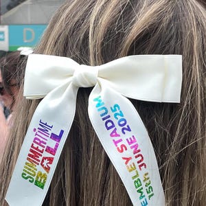 May include: A white hair bow with two long tails featuring colorful text. The left tail reads "SUMMERTIME BALL" in a multi-colored gradient, and the right tail reads "WEMBLEY STADIUM 15th JUNE 2025" in blue and green gradient lettering.